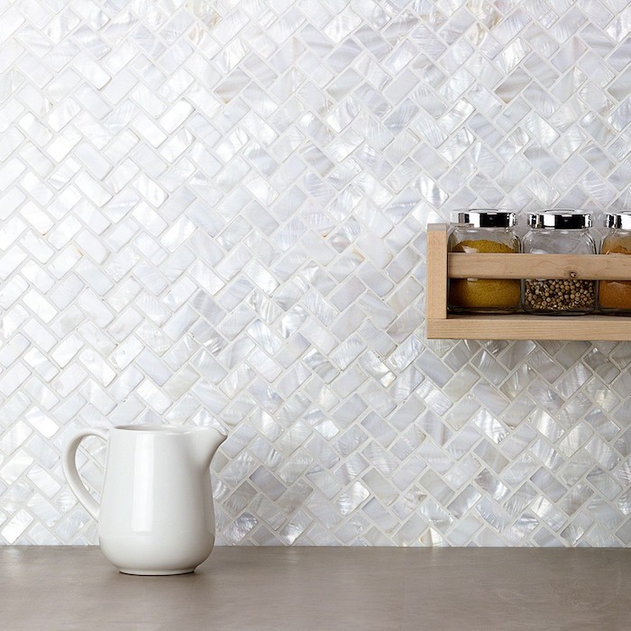 pearlescent white tiles, on a herringbone backsplash, near a beige counter top, with a white milk jug, and a wooden shelf, with condiment jars