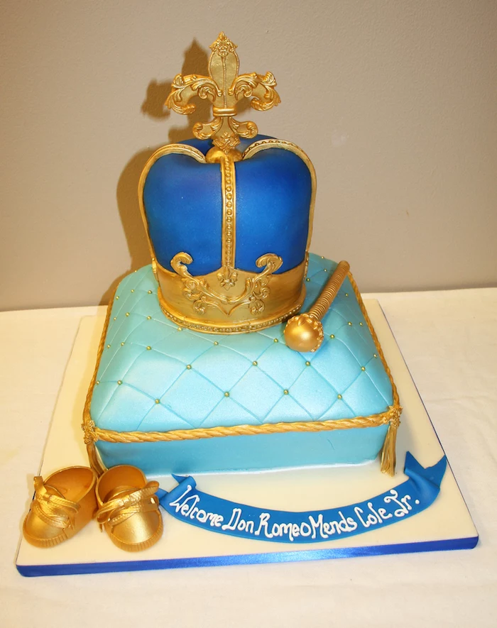 crown made from dark blue, and gold fondant, on top of a cake shaped like a pillow, in light blue and gold, with tiny gold shoes, and a blue ribbon, with white writing