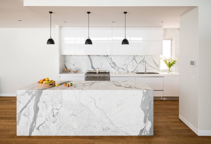 floor covered in brown laminate, under a marble kitchen island, in white and pale grey, with a matching backsplash, smooth white cabinets