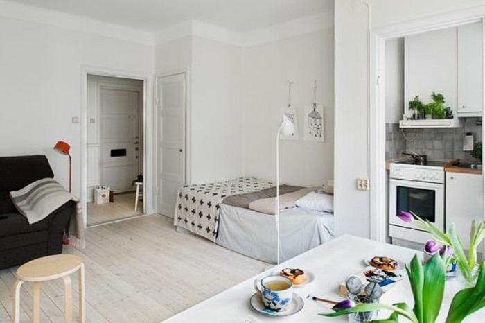 open door in a room with white walls, and a pale beige laminate floor, bed and a black sofa, dining table in white, with flowers and food