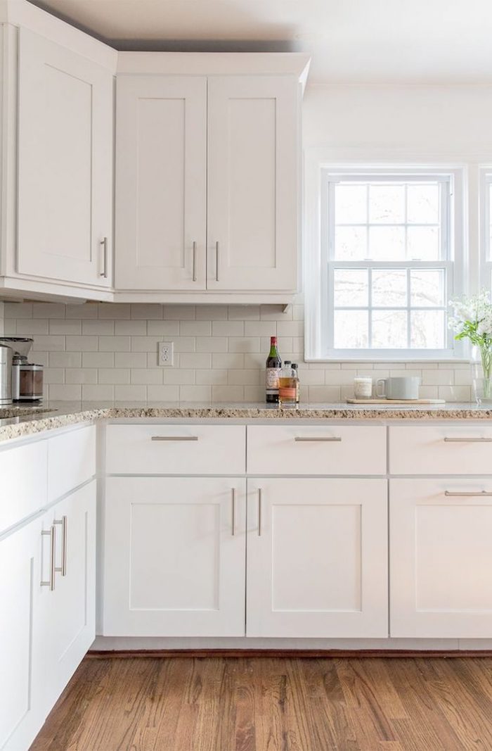 white subway tile pattern, inside a bright kitchen, with brown laminate floor, white cabinets and beige marble counter tops