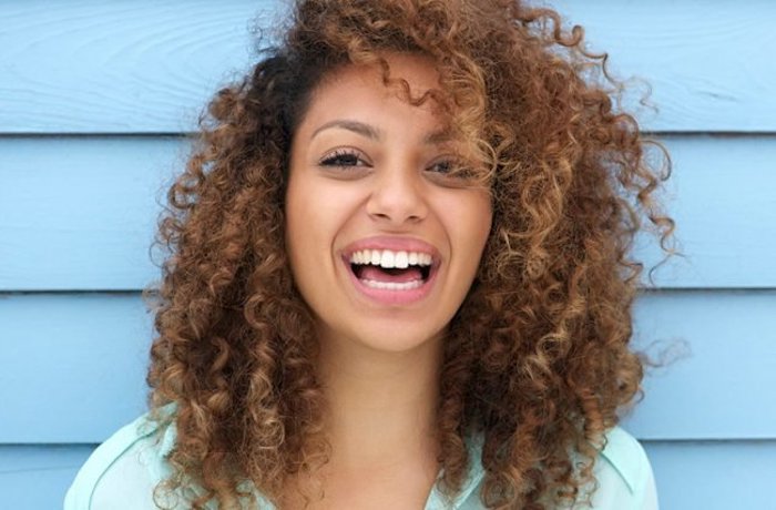 wide smile on the face of a young girl, wearing a pale blue shirt, brunette hair with deep side part, short haircuts for curly hair, ringlets with dark blonde highlights
