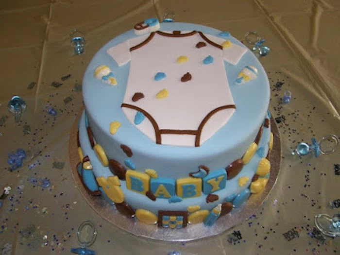 footprints in yellow, brown and blue, decorating a layered light blue cake, topped with a white onesie shape, made from fondant, onesie cake, yellow and blue blocks, spelling out the word baby