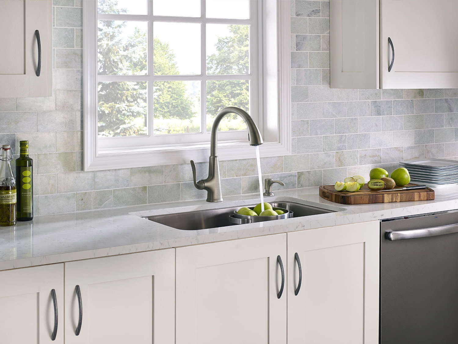 fancy countertops in kitchen in pale tones, with white cupboards, and a metal sink, pale blue brick wall with window, green apples and kiwis