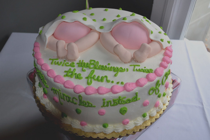 neon green and pale pink frosting, on a white cake, decorated with two sets of baby legs, in pink nappies, under a white blanket with green polka dots