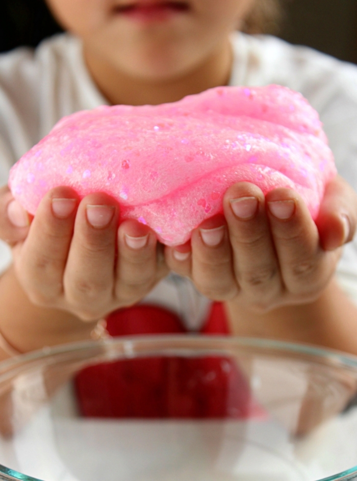 girl holding a pile of fluffy, soft light pink slime, with added pink heart-shaped glitter flakes, over a clear glass mixing bowl