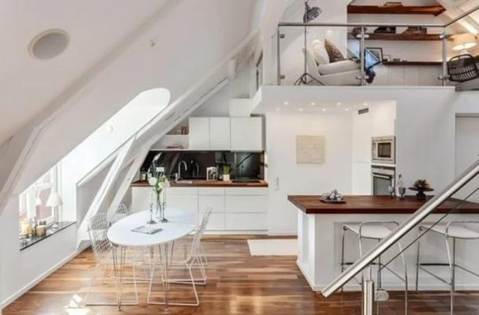 oval dining table in white, and four chairs, inside a loft studio flat, with brown laminate floor, kitchenette and an upper level, with a sitting area