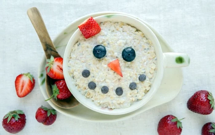 smily face created with fruit, blueberries and strawberry slices, on top of a porridge, inside a large ceramic mug, healthy low calorie breakfast, wooden spoon and whole strawberries
