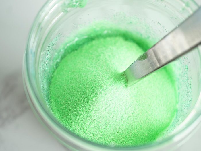 sugar-like green powder, inside a glass container, with a metal, silver colored spoon, fluffy slime recipe, marble patterned surface
