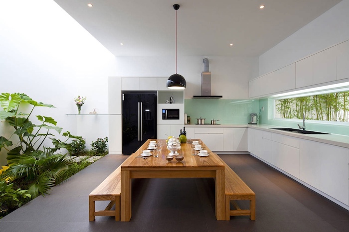 lots of green vegetation, in a large room, with white walls, and a smooth grey floor, long wooden dining table, with two matching benches, turquoise glass backsplash