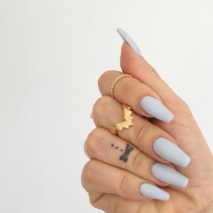 tattoo on the ring finger of a hand, wearing several golden rings, and pale creamy grey nail polish, on matte coffin nails 
