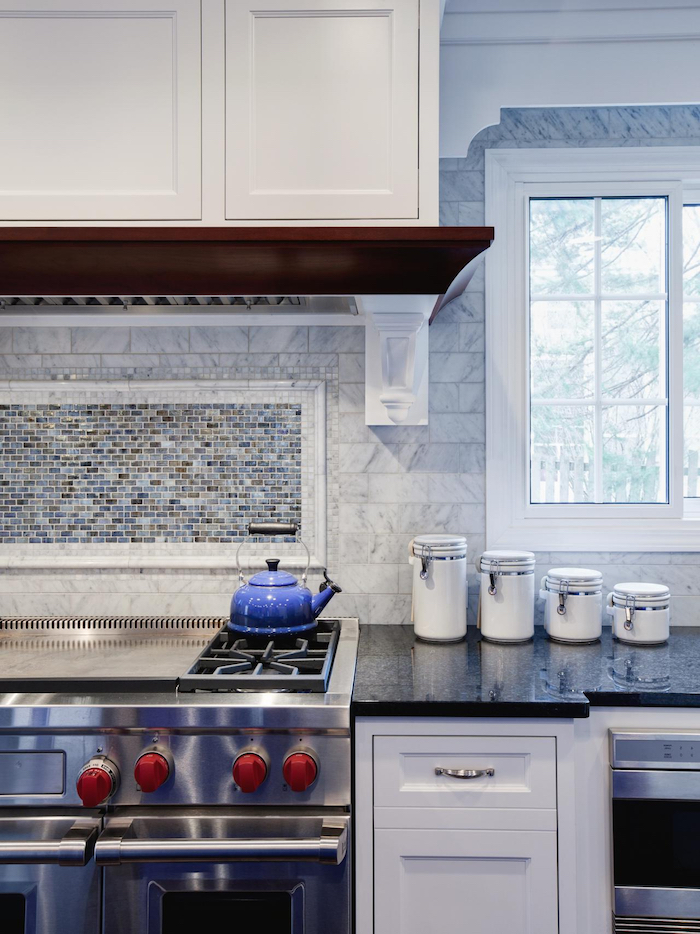 teapot in a vivid blue color, on a metal hob, near a dark counter top, and a pale grey, subway tile kitchen backsplash, with a marble pattern