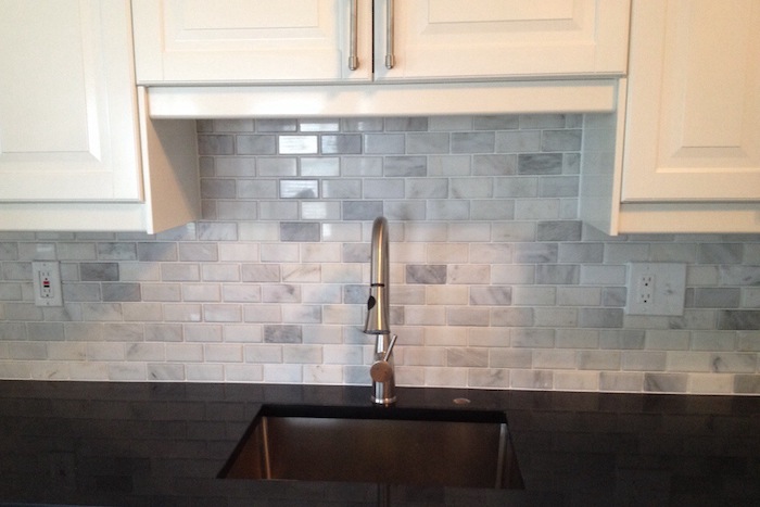 small sink with a metal faucet, in a smooth black counter top, near pale grey subway tile wall, with white kitchen cabinets