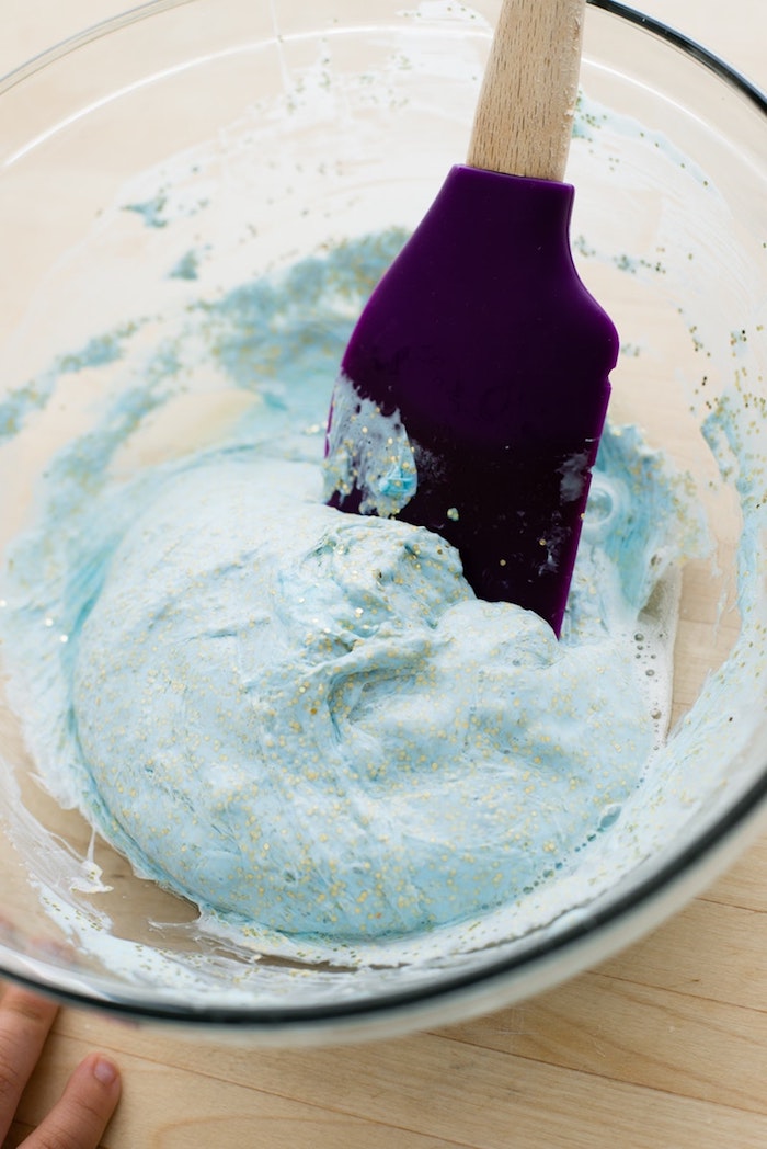 using a plastic spatula in purple, with a wooden handle, to mix blue goop and glitter, fluffy slime recipe, inside a clear glass bowl