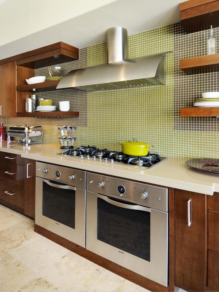 brown wooden cabinets, and a pale cream counter top, in a kitchen with two ovens, light beige tiled floor, and a green and grey mosaic on the wall