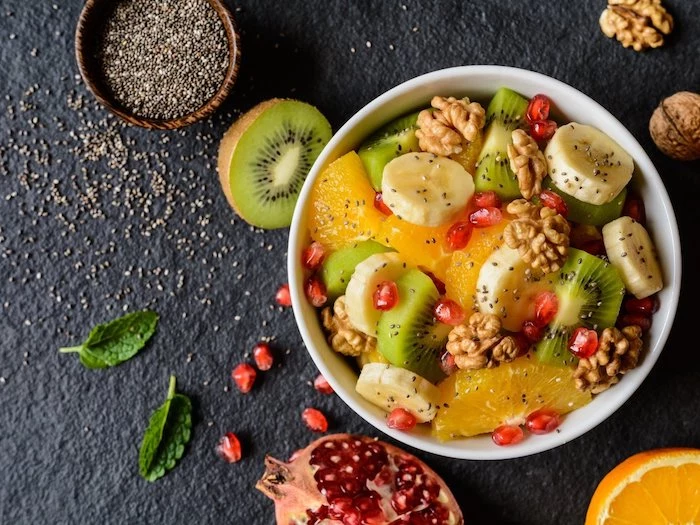 healthy breakfast ideas, white ceramic bowl, filled with slices of fruit, orange and kiwi, banana and pomegranate seeds, and topped with walnuts