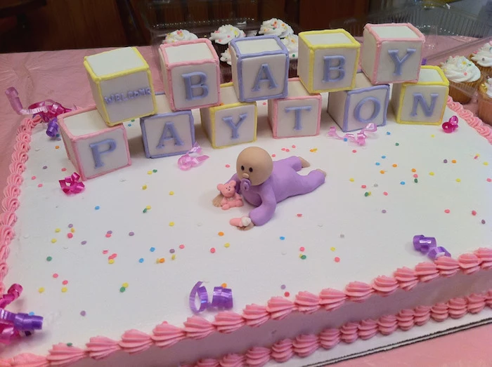 confetti in different colors, strewn on a white cake, with pink frosting, baby shower sheet cakes, decorated with a fondant baby figurine, in a purple onesie, and several alphabet blocks, in different colors, spelling out a festive message