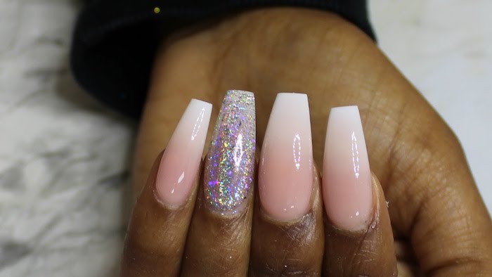 brown hand with long, coffin acrylic nails, painted in milky pink and white, with ombre-like effect, the ring finger nail is covered in iridescent glitter