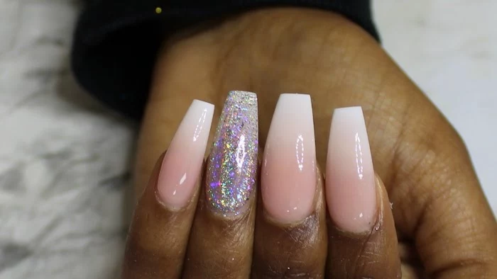 brown hand with long, coffin acrylic nails, painted in milky pink and white, with ombre-like effect, the ring finger nail is covered in iridescent glitter