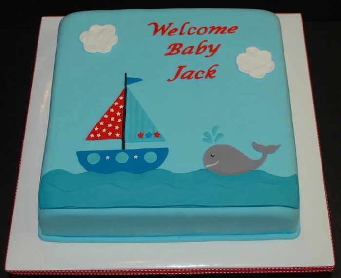 sailing boat in blue, red and turquoise, decorated with little stars, on a light blue square cake, nautical baby shower cakes, with a little grey whale, and a festive message in red 