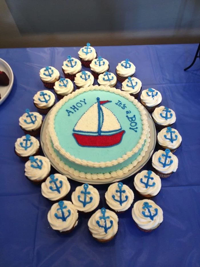 ahoy it's a boy, written in blue frosting, on a round pale turquoise cake, with a red and white drawing of a sailing boat, nautical baby shower cakes, surrounded by cupcakes topped with anchors
