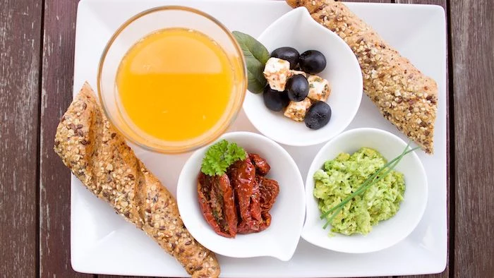 mediterranean breakfast on a tray, with large glass of orange juice, two pieces of bread, and three small white dishes, containing guacamole and sun-dried tomatoes, olives and marinated cheese