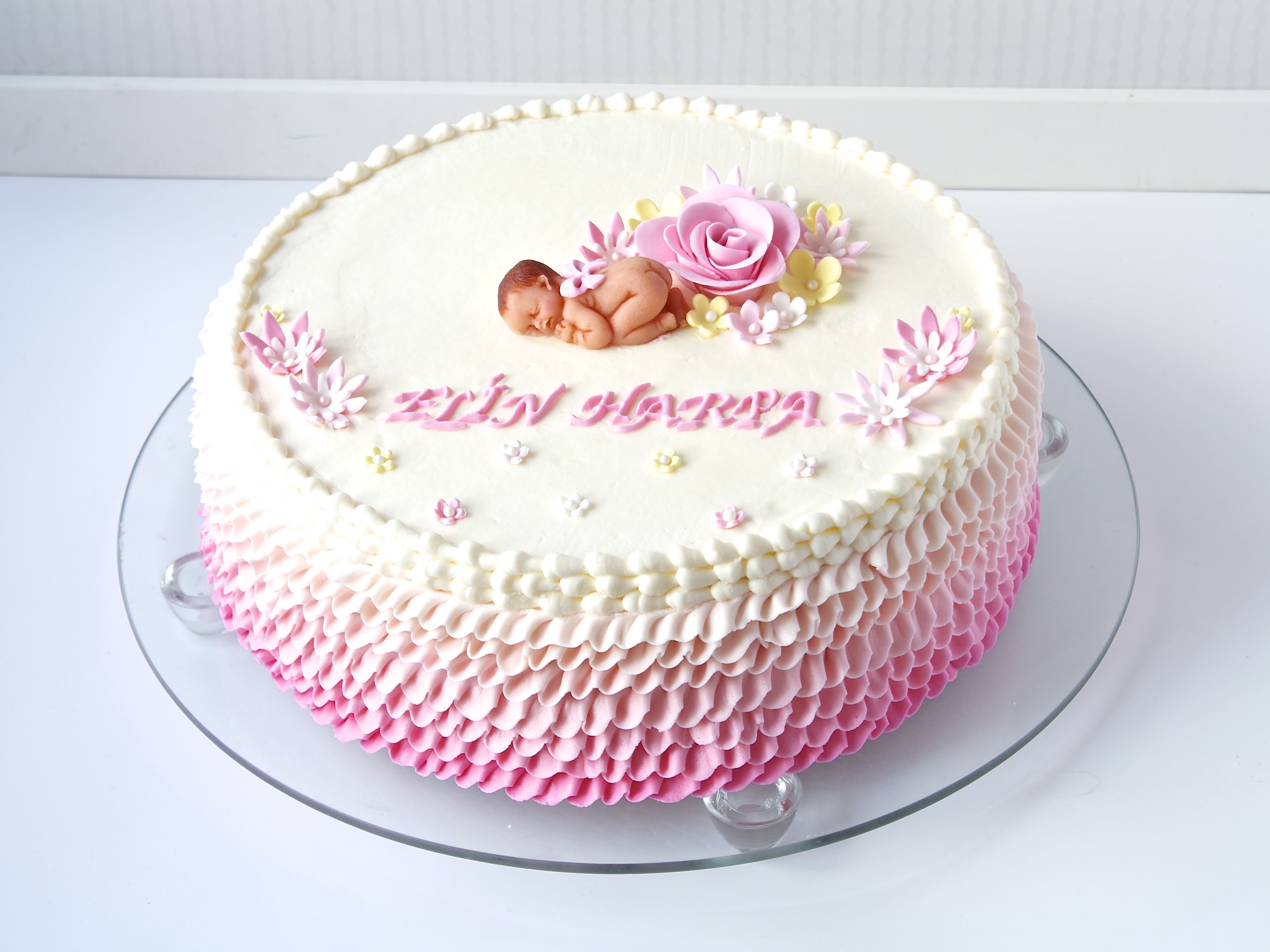 frills in white and different shades of pink, made from frosting, decorating a cake, topped with a tiny, realistic baby figurine, and flowers made from fondant, baby shower cakes for girls