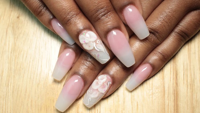 pale wooden surface, under two brown hands, with long nails, painted in ombre-like pink and white, ring finger nail is decorated with white acrylic flowers, ballerina nail shape