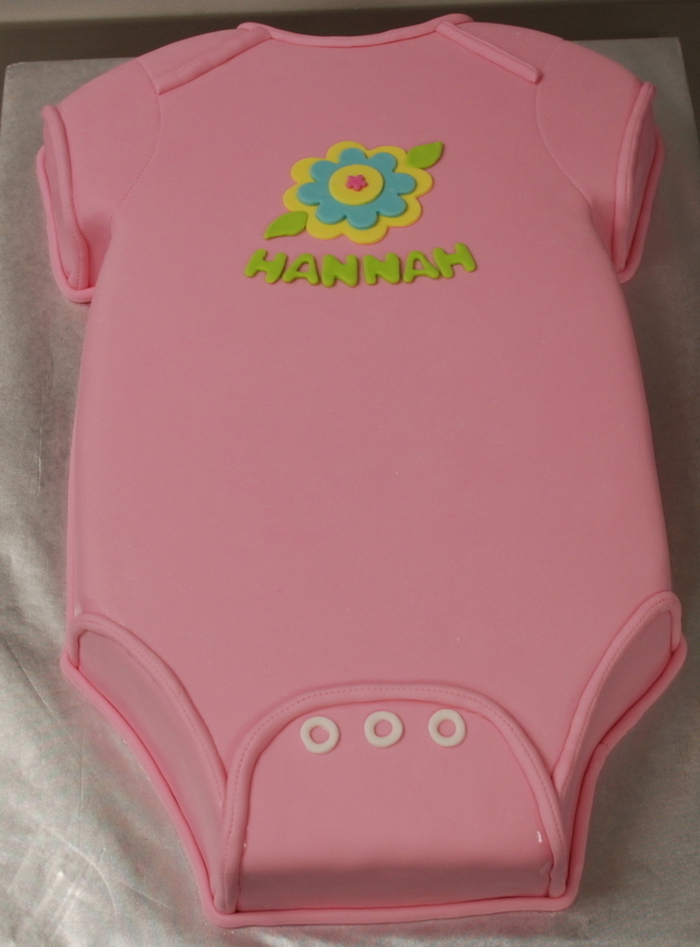 onesie cake in baby pink, decorated with a fondant flower, in yellow and turquoise, green and pink, with the name hannah