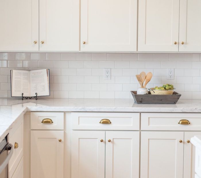 book on a black metal stand, placed on a white marble counter, in kitchen with white cabinets, white subway tile pattern on the wall, little grey wooden box wth utensils 