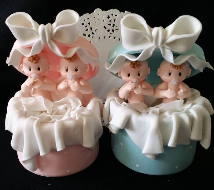 twin baby shower cakes, cake toppers in white and pink, and white and blue, shaped like opened gift boxes, with white bows, with a set of two baby figurines in each