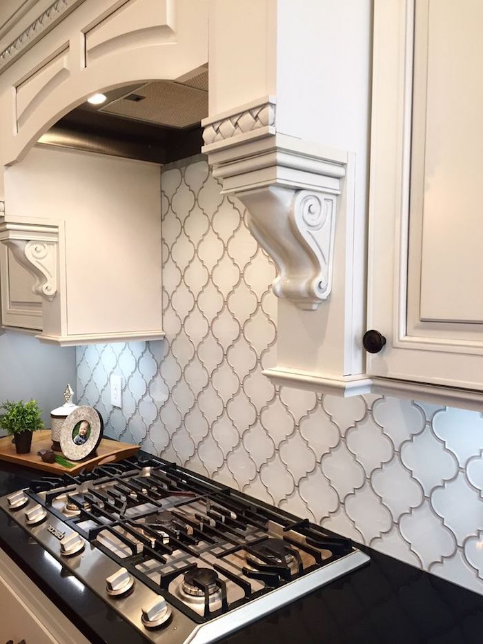 arabesque backsplash in white, with grey joints, near a black counter top, with an inbuilt hob, and white cabinets, with ornamental details