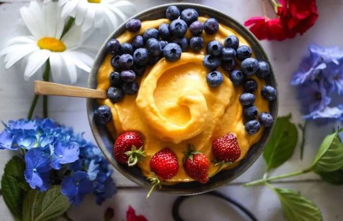 daisies and other flowers in blue and red, placed around a bowl, containing orange mush, low calorie breakfast, garnished with blueberries and strawberries