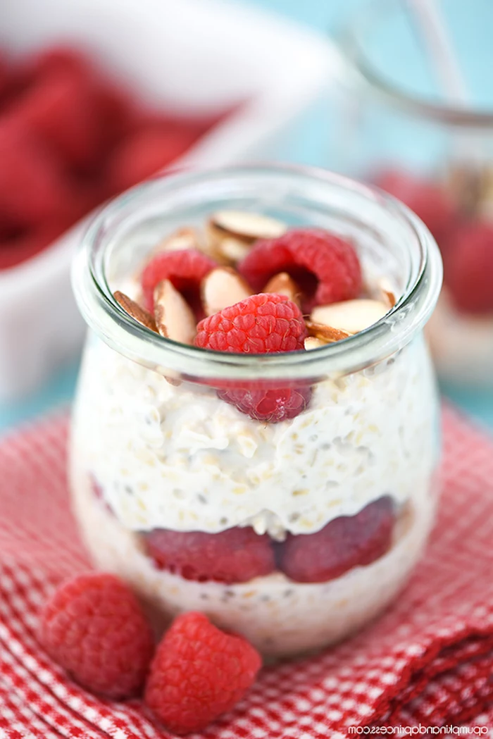 small pot made of clear glass, containing yoghurt and rolled oats, with raspberries and almonds, easy breakfast recipes, placed on a red and white checkered napkin