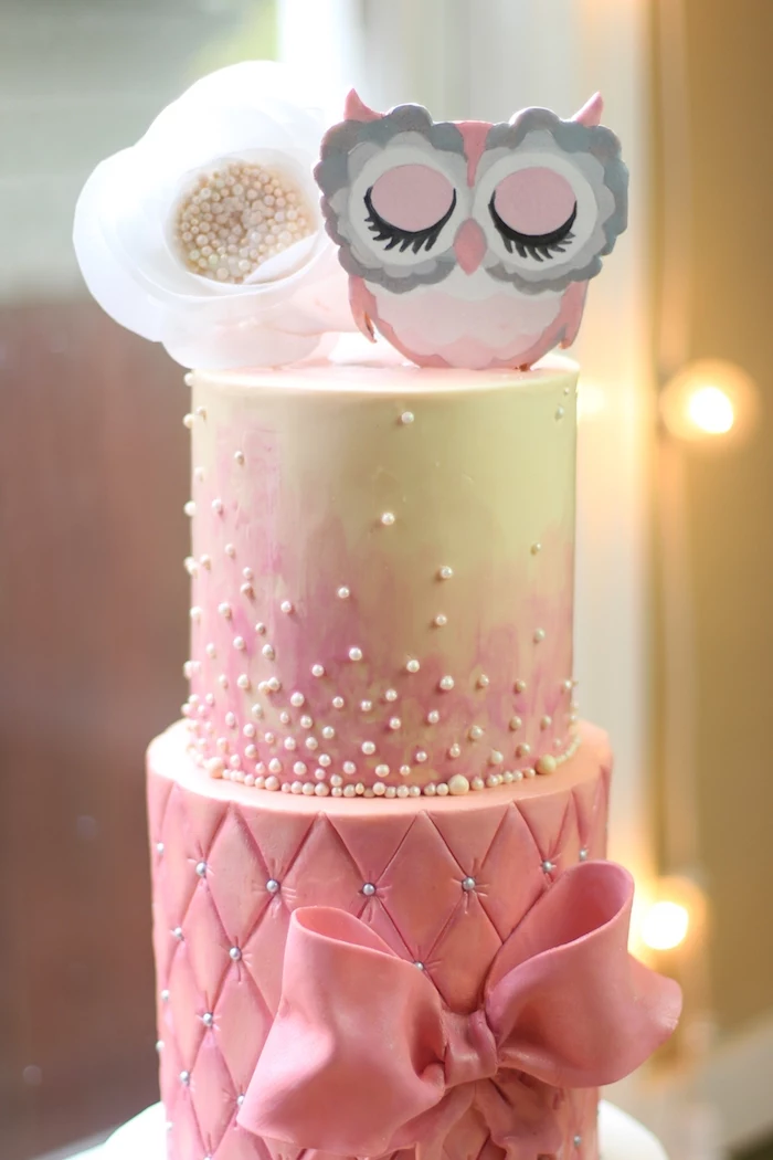 owl baby shower cake, in pale pink, and light cream, decorated with a large fondant bow, white edible pearls, a large faux white flower, and a pink owl figurine