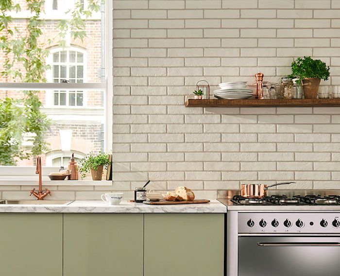 khaki green kitchen cabinets, pale and smooth, in a room with a shiny metal oven, tiled wall in cream, wooden shelf with utensils