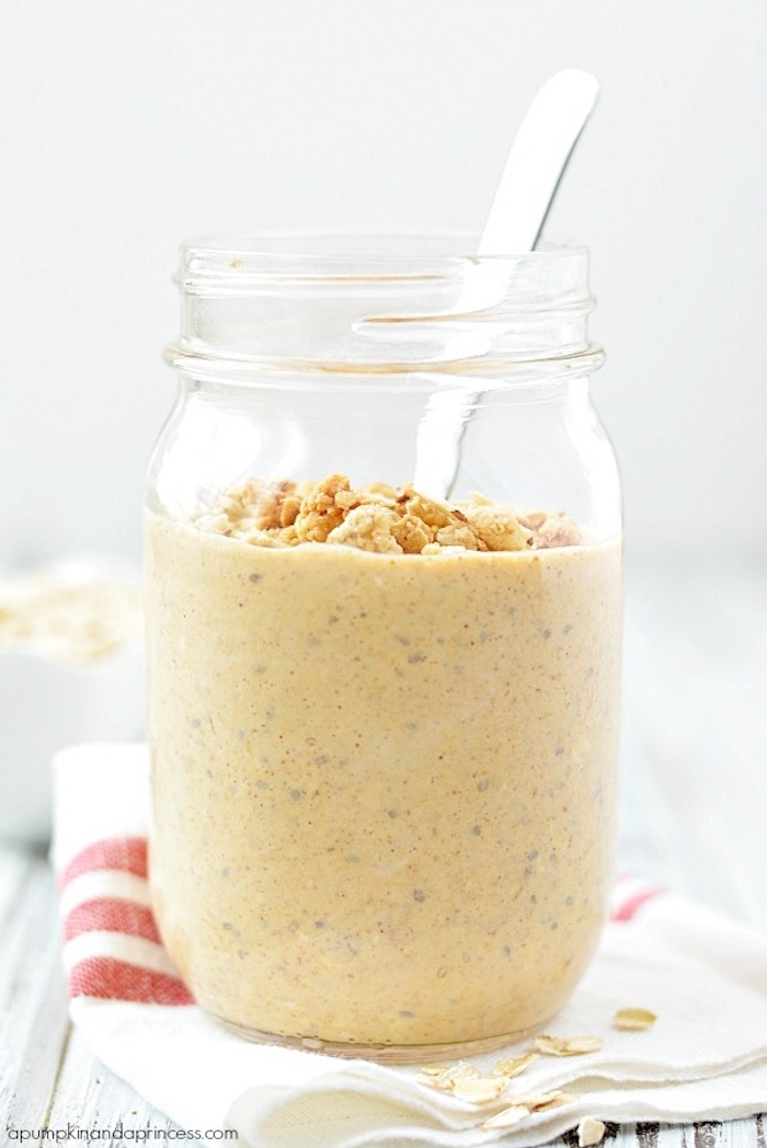 metal spoon inside a large clear jar, containing a pale orange porridge, topped with rolled oats, and crunchy granola, simple breakfast ideas, placed on a folded napkin