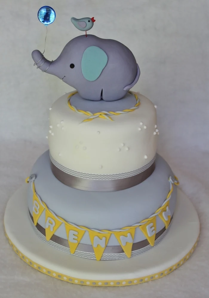 balloon in metallic blue, held by a grey elephant figurine, with turquoise ears, placed on top of a layered cake, with pale grey and yellow details