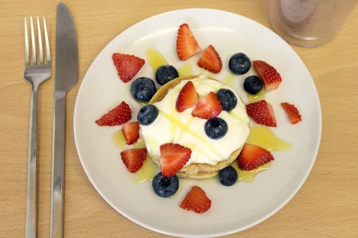 honey drizzled over a stack of small pancakes, covered in yoghurt, and garnished with pieces of fresh strawberry and blueberries