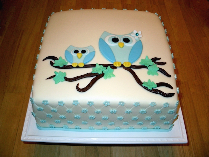 mother and baby owl, made from blue fondant, perched on a brown branch with green leaves, on top of a white, square-shaped cake, owl baby shower cake, with pale blue details