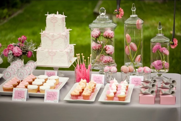 baby shower cakes for girls, a selection of different cupcakes and pastries, in pink and white, on a grey table, near a three-layered cake, and several flower vases