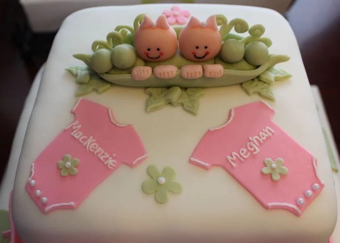 mackenzie and meghan, written in white, on two pink onesies, decorating a smooth white, square-shaped cake, twin baby shower cakes, topped with a green pea pod, containing two baby figurines
