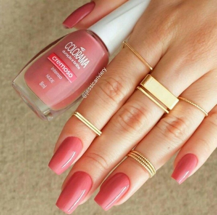 square tips on oval nails, painted in peach pink nail polish, on a hand with several square and round gold rings, holding a bottle of nail polish