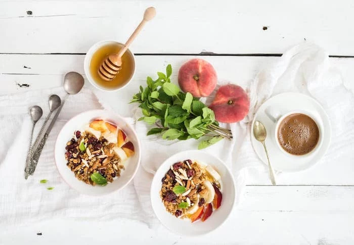 sprigs of mint and two peaches, near a bowl of honey, and a cup of coffee, healthy low calorie breakfast, two bowls of muesli, covered with fruit