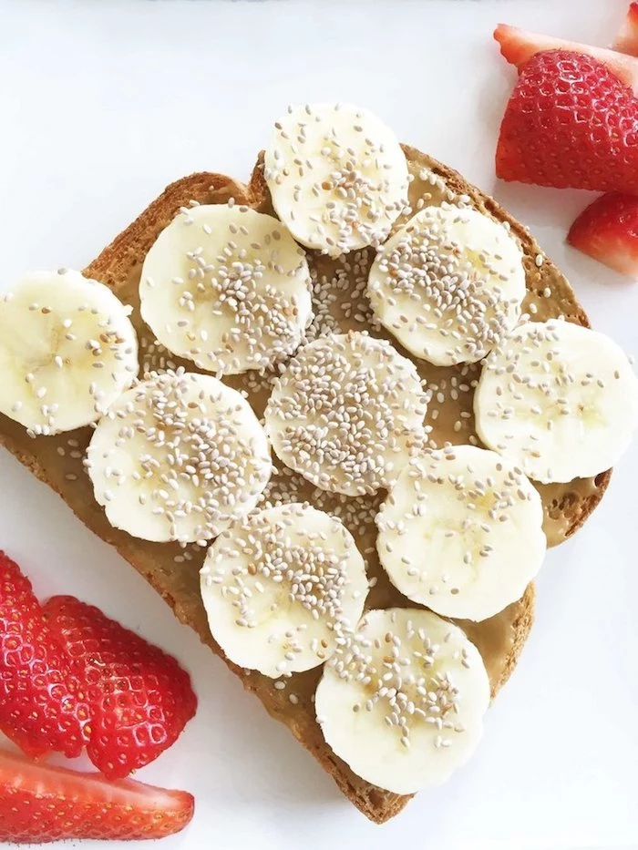 sesame seeds on slices of banana, on top of a piece of toast, smeared with peanut butter, easy breakfast recipes, next to several pieces of strawberry