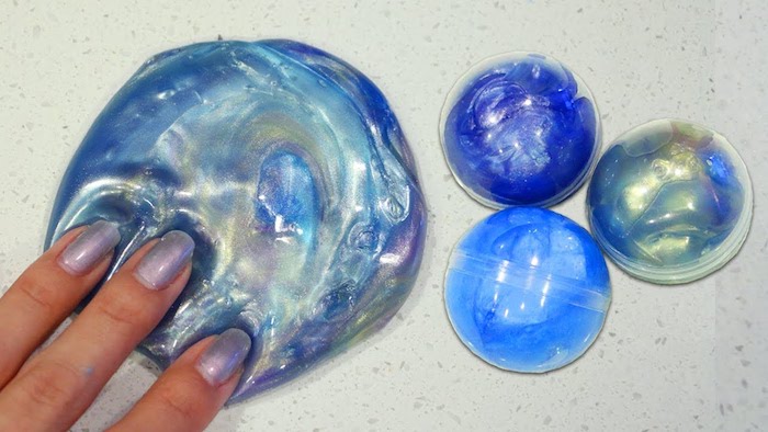 three fingers pressing on a round piece of slime, in several shades of blue, with a pearly sheen, three plastic orbs, containing more blue slime nearby
