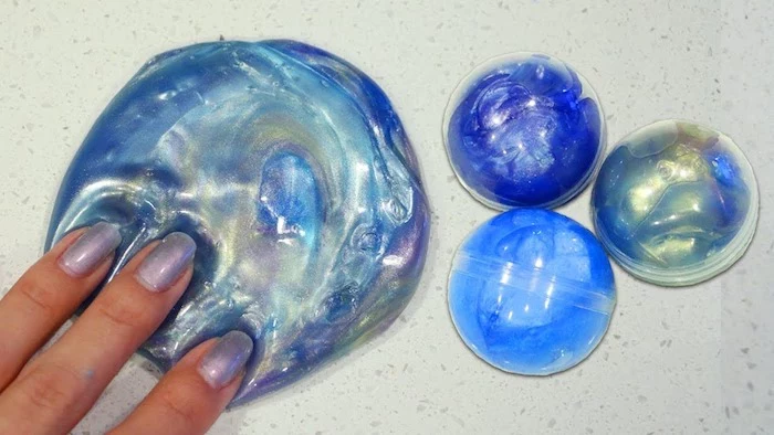 three fingers pressing on a round piece of slime, in several shades of blue, with a pearly sheen, three plastic orbs, containing more blue slime nearby