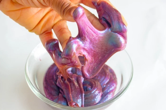 metallic-effect galaxy slime in pink, with hints of blue and orange, how to make slime with shaving cream, stuck to the fingers of a hand, holding it over a glass bowl with more slime