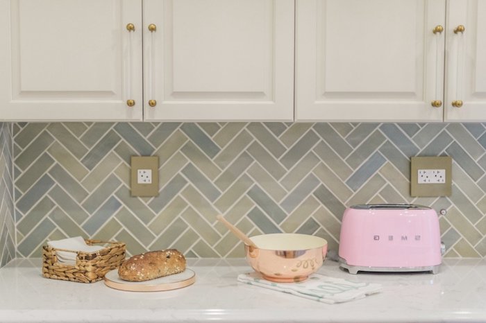 rose gold metallic bowl, with a wooden spoon, near a candy pink toaster, on a light counter top, with a herringbone backsplash, in different tones of grey 