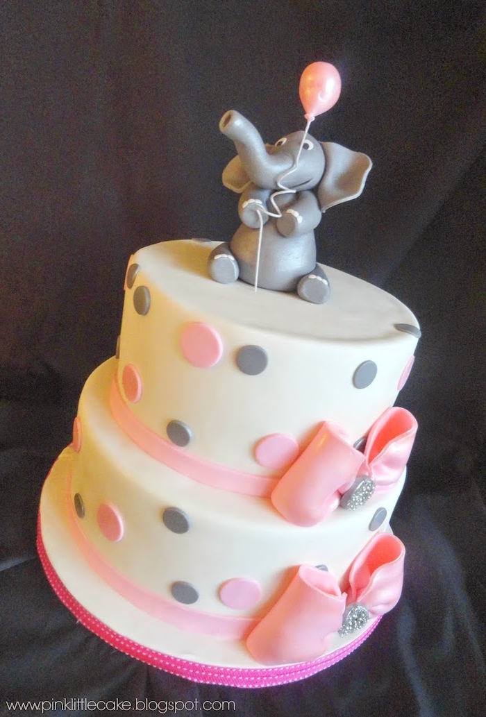 grey fondant figurine of an elephant, holding a pale pink balloon, on top of a layered cake, covered with white frosting, and decorated with pale pink, and light grey polka dots, and two pink ribbons with bows